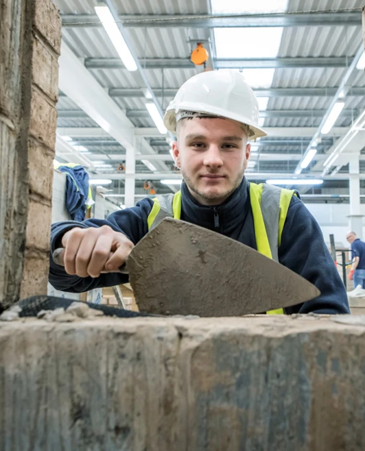 A brickwork student wearing a safety helmet and high-visibility vest using a trowel to apply mortar, viewed through an opening in a brick structure. A brickwork student wearing a safety helmet and high-visibility vest using a trowel to apply mortar, viewed through an opening in a brick structure.